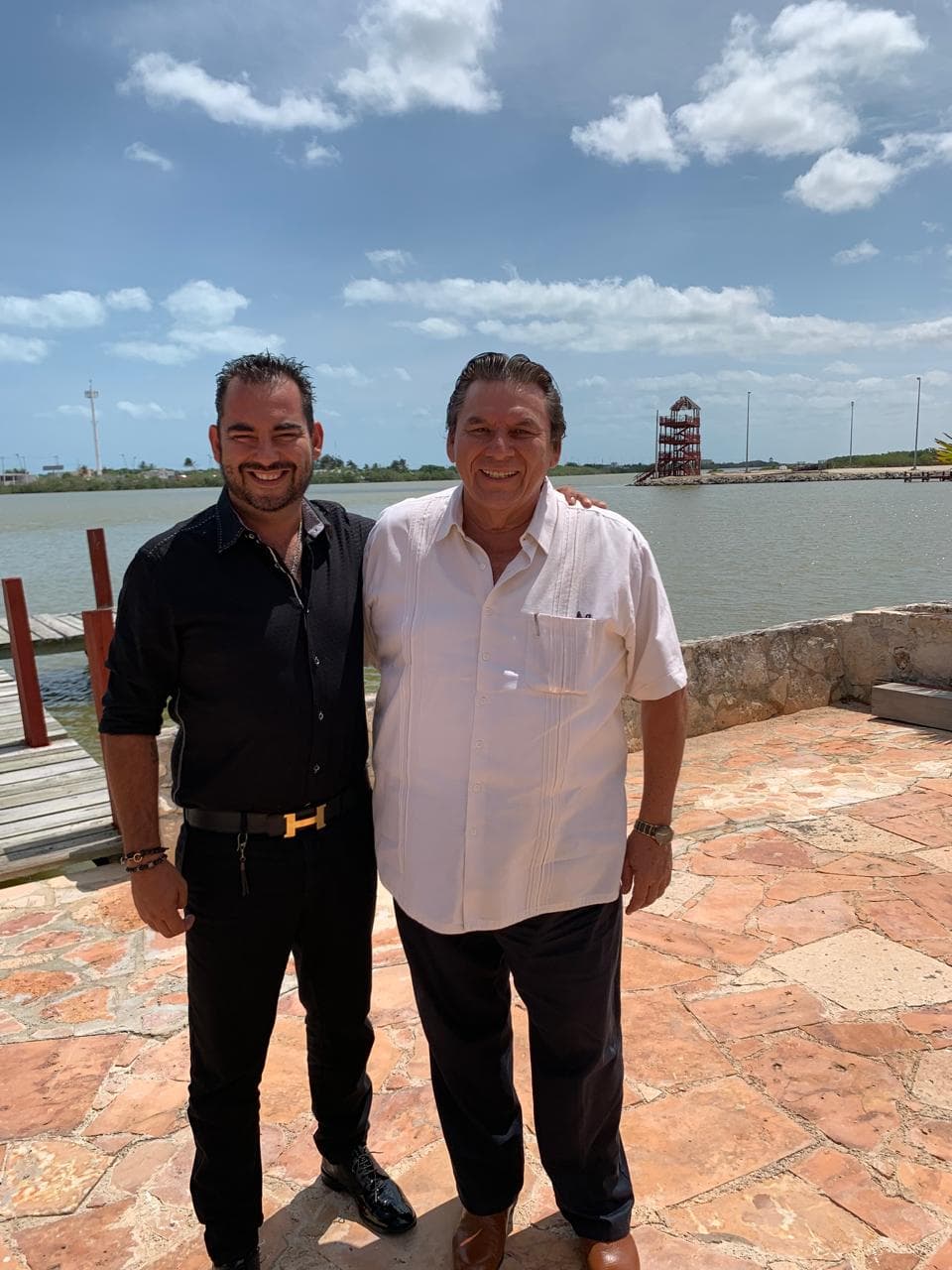 Julio Ortiz Mena, con camisa negra, sonríe junto a un hombre con guayabera blanca en una terraza de piedra frente al agua, bajo un cielo azul.