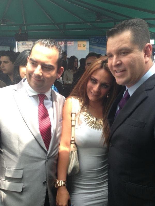 Julio Ortiz Mena, con traje gris claro y corbata roja, posa junto a una mujer y un hombre durante un evento bajo una carpa verde.
