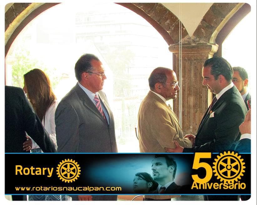 Julio Ortiz Mena saluda a un hombre de saco claro bajo arcos de cantera durante una actividad del Club Rotario de Naucalpan, con la franja original «Rotary · 50 Aniversario».