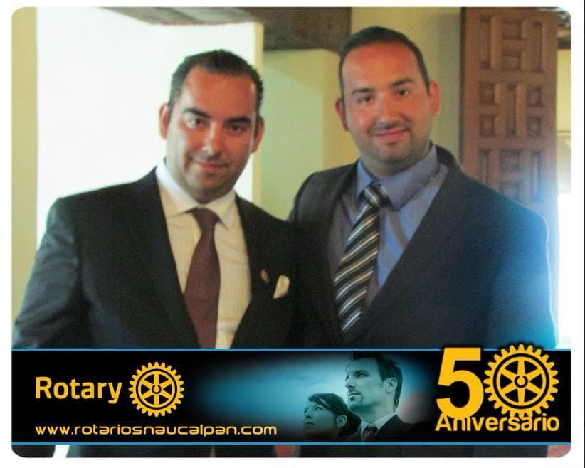 Julio Ortiz Mena, con corbata vino y pañuelo blanco, posa junto a un hombre de traje oscuro y corbata a rayas, con la franja original «Rotary · 50 Aniversario».
