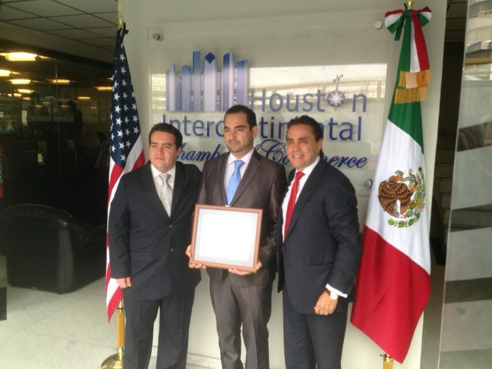 Julio Ortiz Mena sostiene un marco junto a dos hombres de traje, entre banderas de México y Estados Unidos, frente a una señalización institucional.